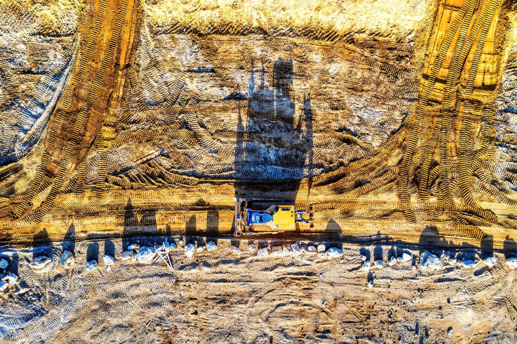 Aerial view of a tracked dozer at the floor of a wide excavation, ringed by track marks cut into ochre earth.