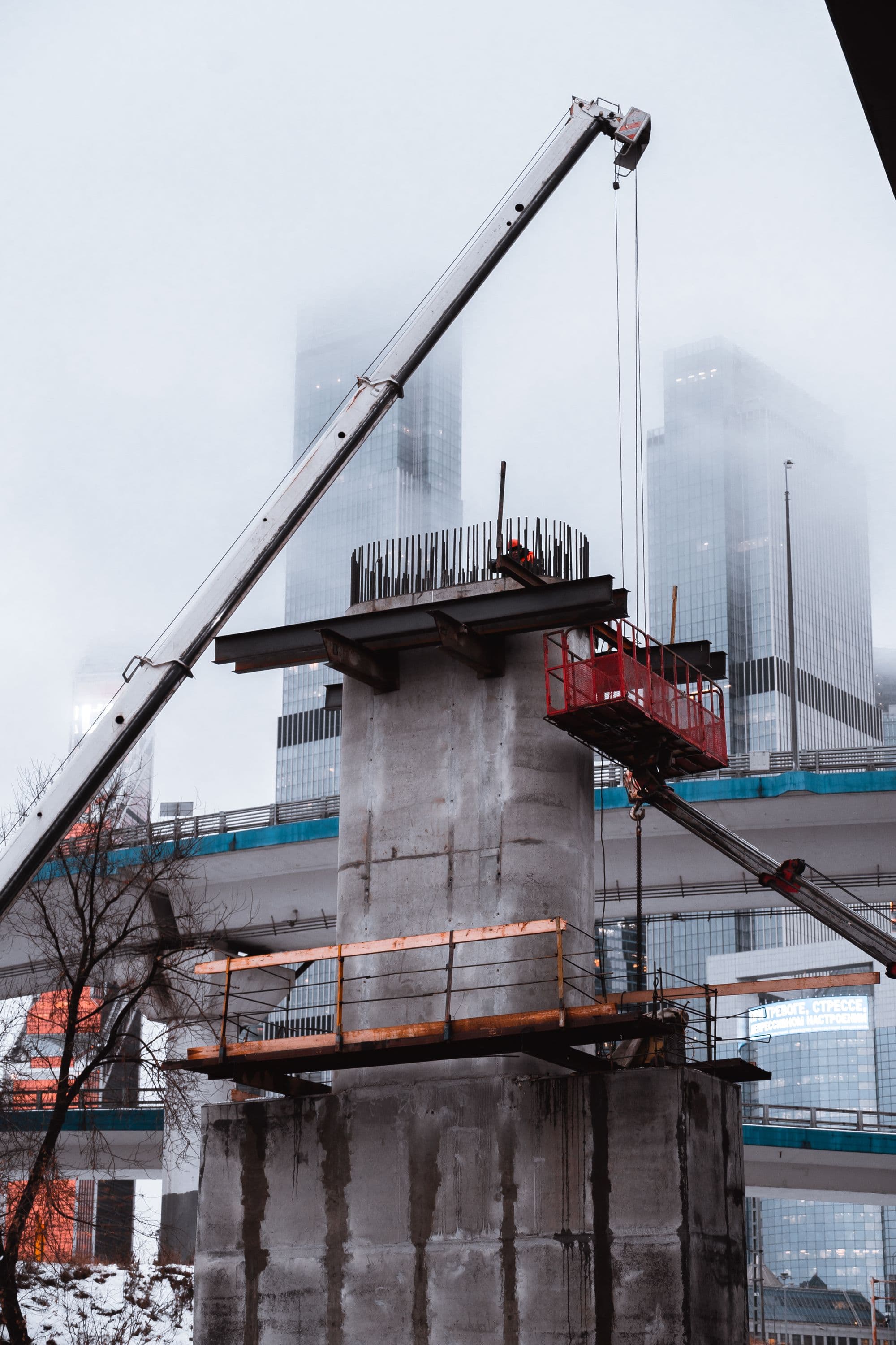A mobile crane setting steel girders onto a concrete bridge pier on a foggy day, city towers fading behind the mist.