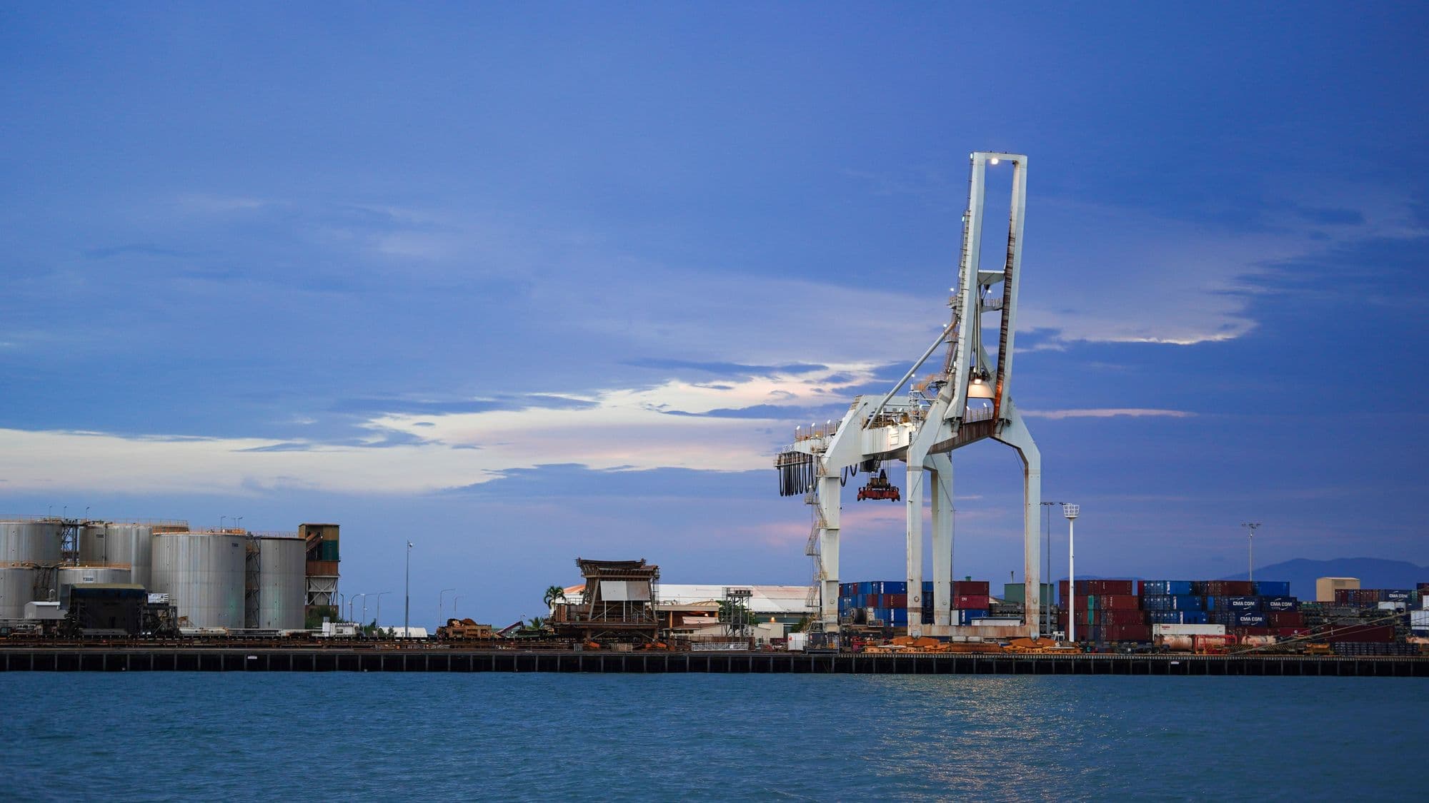 A ship-to-shore gantry crane, stacked containers and storage tanks along a port quay at dusk, blue sky reflected on the water.